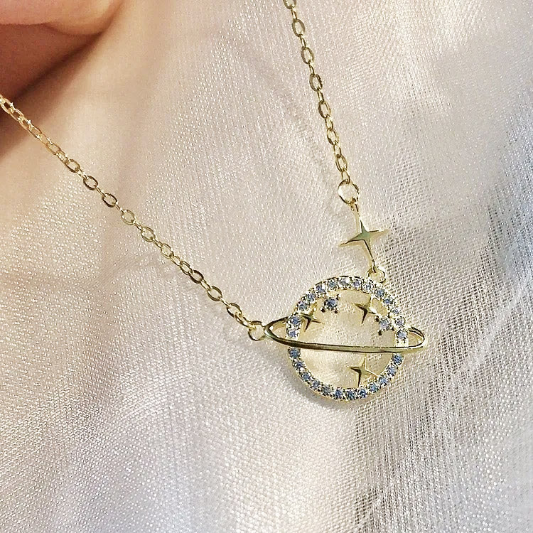 A close-up of a gold-plated 925 sterling silver necklace featuring a minimalist Saturn planet pendant with a central white CZ stone and surrounding clear stones.