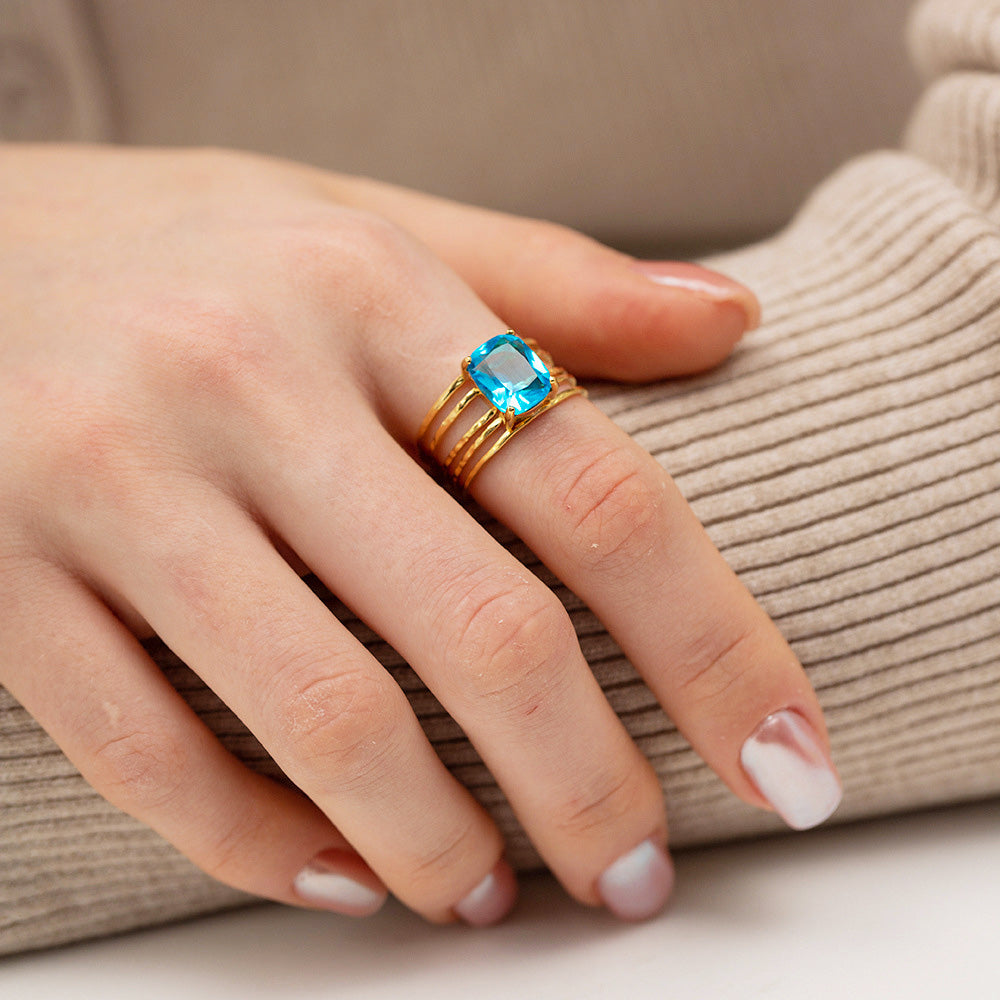 A close-up of a woman's hand wearing the multilayer stainless steel ring with a gold finish, featuring a prominent rectangular blue zirconia gemstone. The ring has a unique stacked band design.