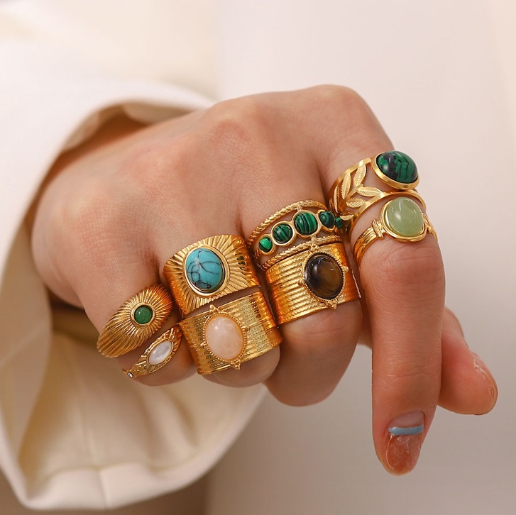 A collection of ornate gold-plated stainless steel rings with various semi-precious gemstones, including green, blue, and white stones, displayed on a hand model.