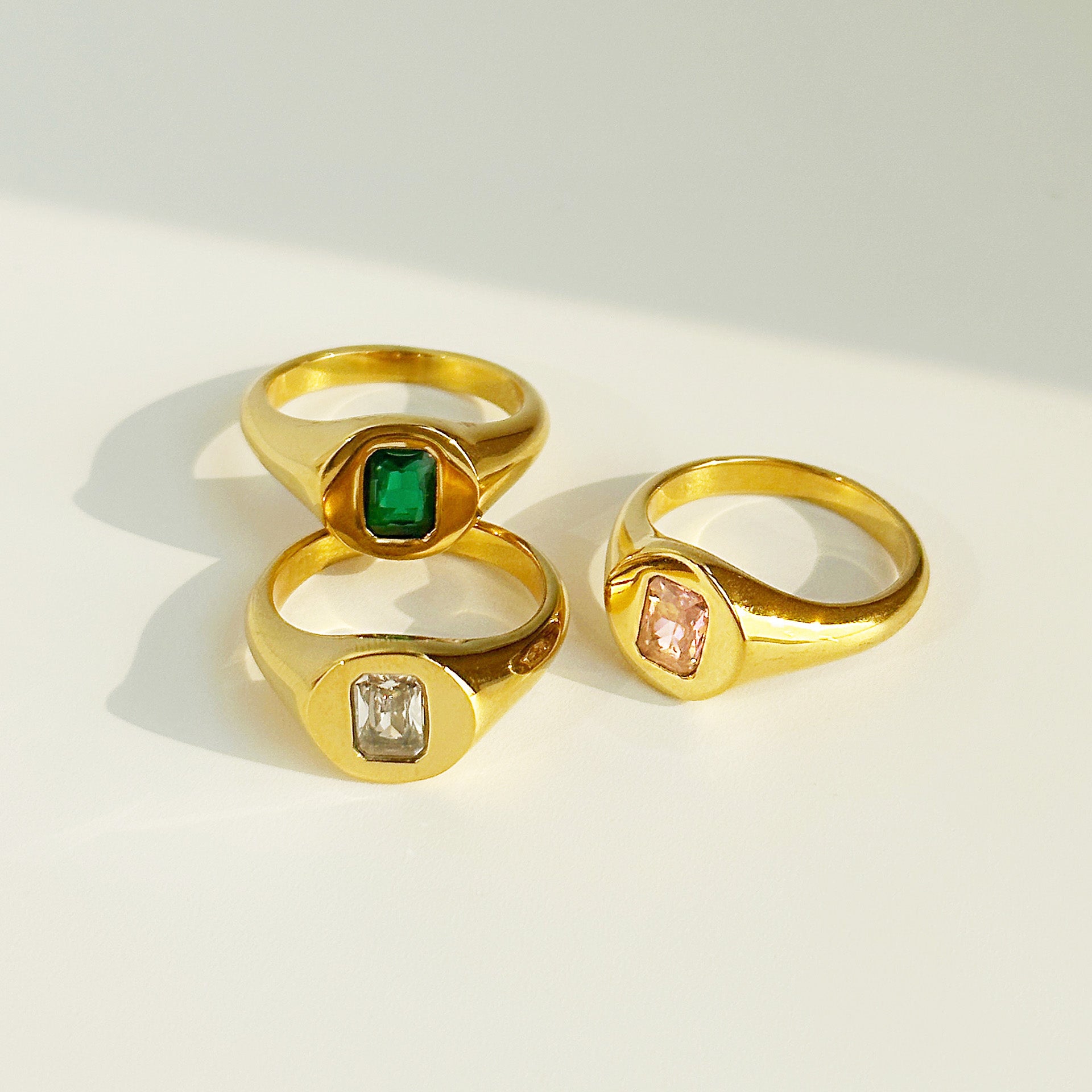 A set of three gold-plated stainless steel signet rings, each featuring a square-cut CZ stone in green, clear, and pink.