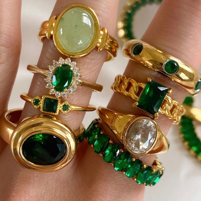 A collection of wholesale stainless steel emerald rings with gold plating, showcasing various designs with green and clear gemstones.