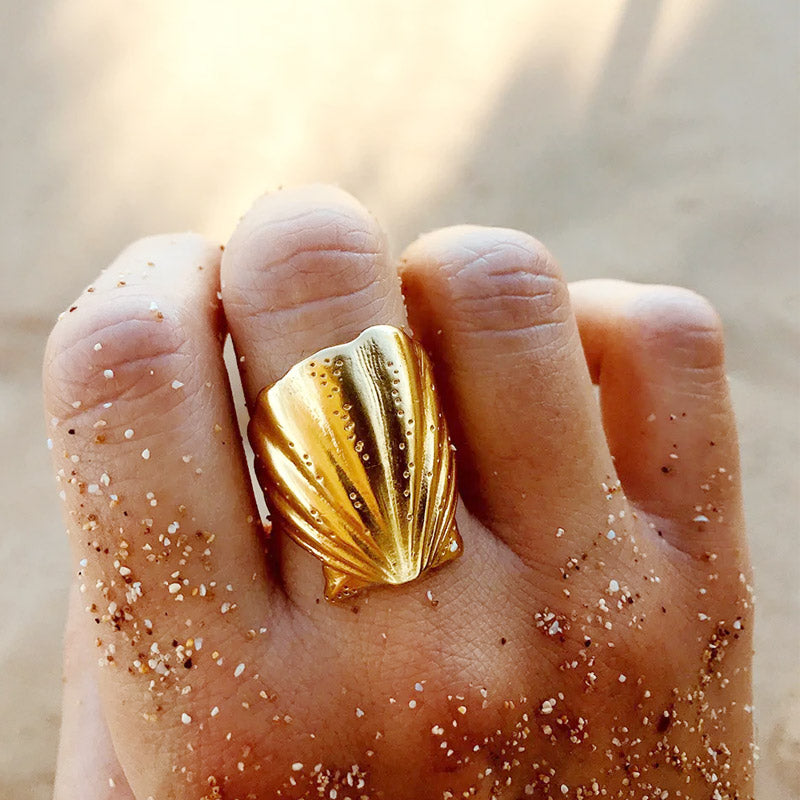 A model's hand wearing a gold-tone, chunky, shell-shaped open ring from the tarnish-free stainless steel beach jewelry set, resting on a sandy surface.