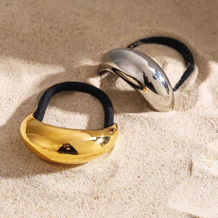 A pair of stainless steel hair tie cuffs, one in a polished gold finish and the other in silver, displayed on a sandy surface.