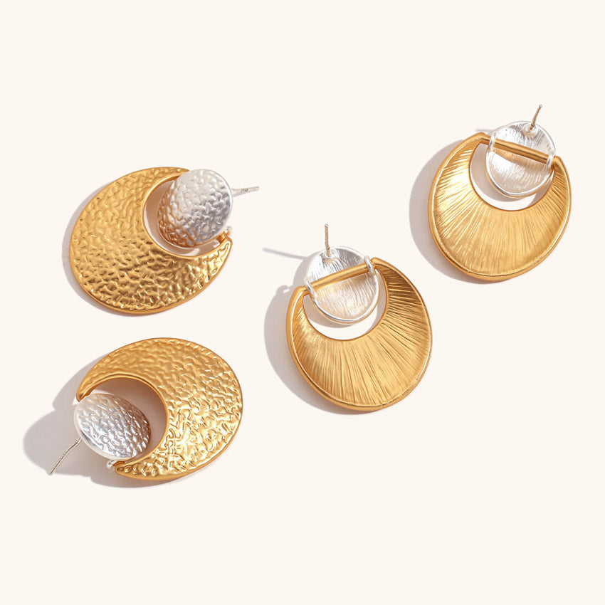 A pair of geometric stainless steel earrings with a two-tone design, featuring a hammered gold crescent moon attached to a silver circle stud, shown on a white background.