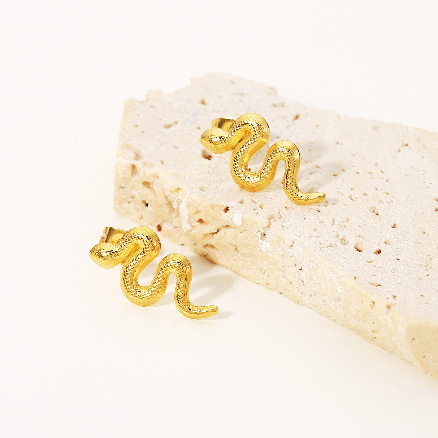 A pair of gold-plated stainless steel snake stud earrings displayed on a textured beige stone, highlighting their unique distorted design.
