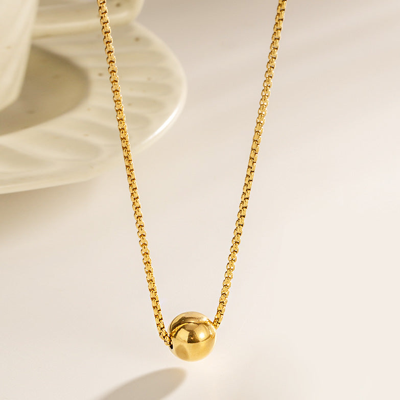 A close-up of a minimalist gold-plated stainless steel necklace featuring a smooth, spherical ball pendant on a delicate pull-box chain.