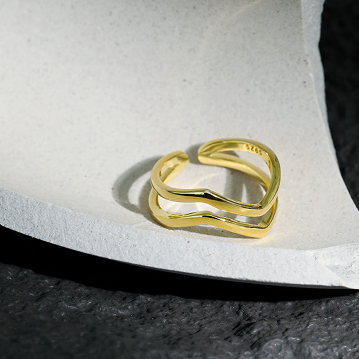 A close-up of a single 18K gold-plated 925 sterling silver wave ring, showcasing its minimalist, flowing design on a textured surface.