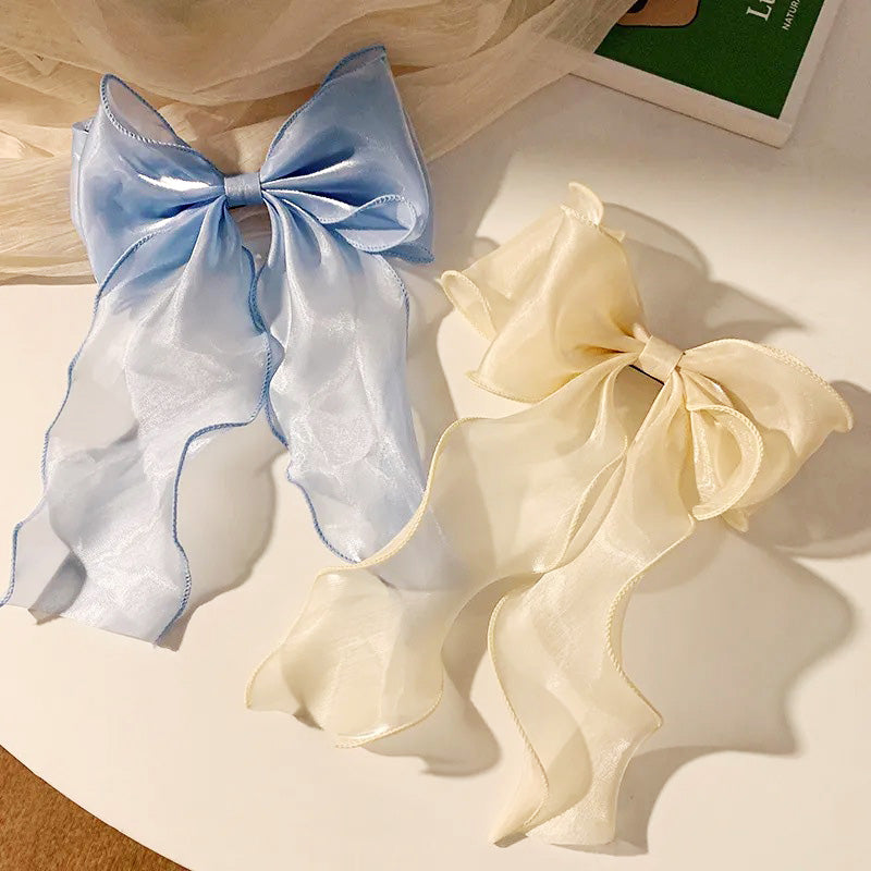 A pair of pearlescent ribbon hair bows, one in light blue and one in cream, showcasing their shiny fabric and elegant design.