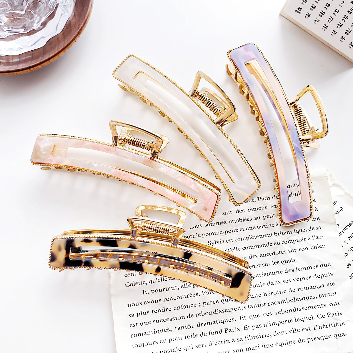 A collection of women's acetate hair claws in various colors like pink, purple, white, and tortoiseshell, all featuring a stylish gold metal finish.