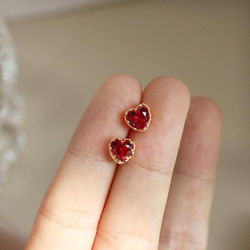 A close-up of delicate 925 sterling silver stud earrings, featuring a gold-plated red heart design, held between fingers to show scale.