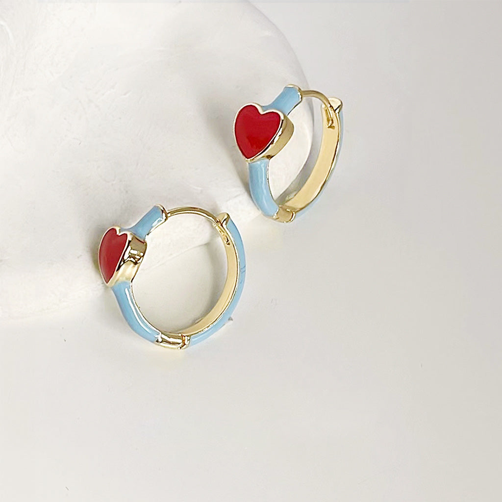 A pair of 14k gold plated huggie hoop earrings with light blue enamel bands and a vibrant red enamel heart at the top, shown against a white background.
