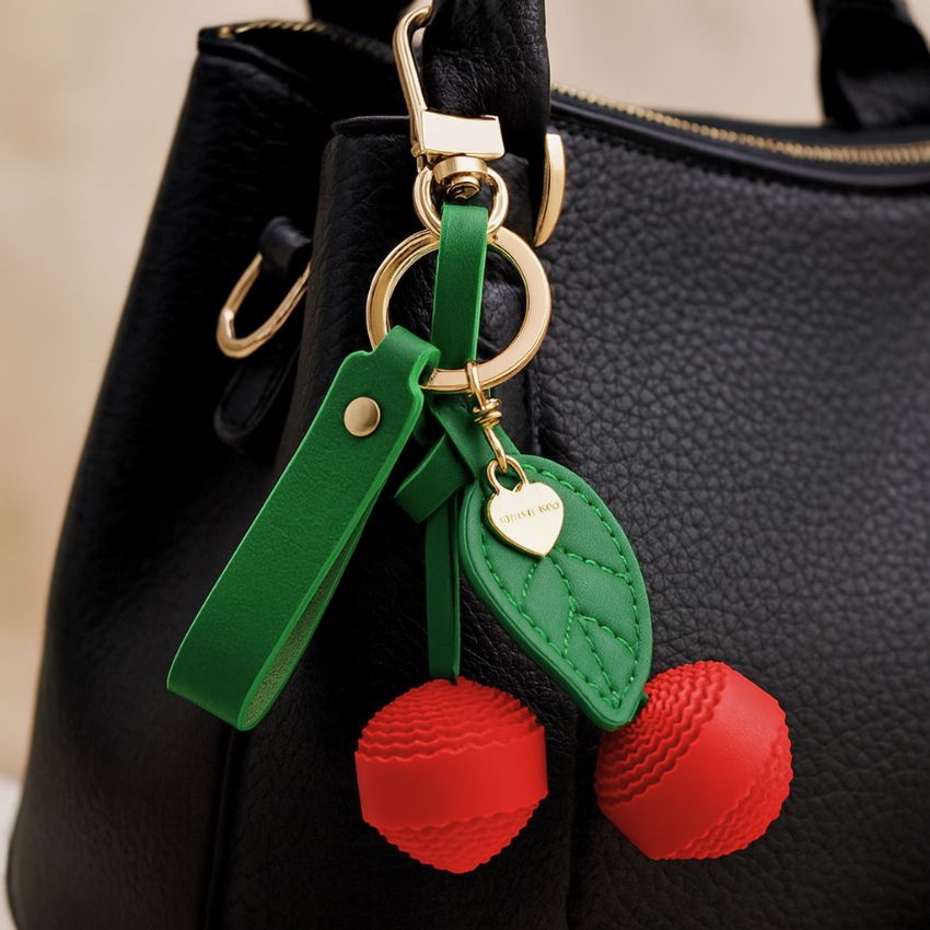 A stylish red PU leather cherry keychain with a green leaf charm attached to the handle of a classic black handbag, showcasing its use as a cute bag accessory.