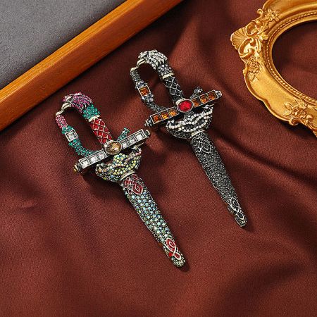 Two variations of the bejeweled sword brooch, one with blue and one with orange crystals, displayed on a rich brown fabric.