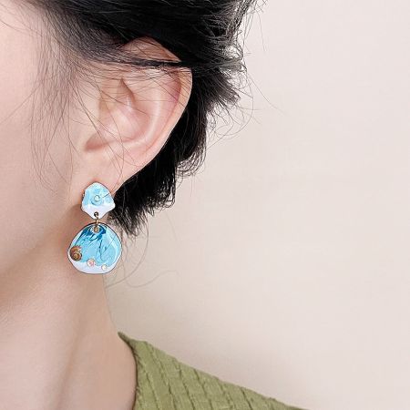 Close-up of two-part drop earrings with a blue and white ocean wave enamel pattern and gold rims.