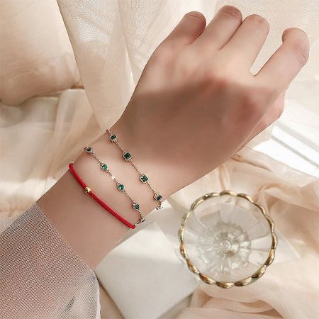 A layered look on a wrist, featuring two gold chain bracelets with green gems and a simple red cord bracelet.