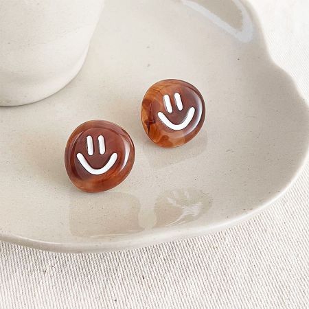 Orecchini a bottone in acrilico marrone smiley con smalto bianco (5 paia) - Un paio di orecchini a bottone in acrilico marrone con design smiley bianco, esposti su una superficie bianca pulita.