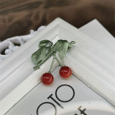 The cherry brooch displayed against a textured white background, emphasizing its vibrant colors.
