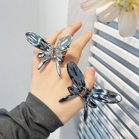 A hand holding two butterfly hair clips, one in silver and one in a dark gun-black finish.