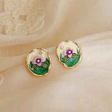 Detailed view of the gold-rimmed floral earrings with pink flowers and green leaves on a cream fabric background.