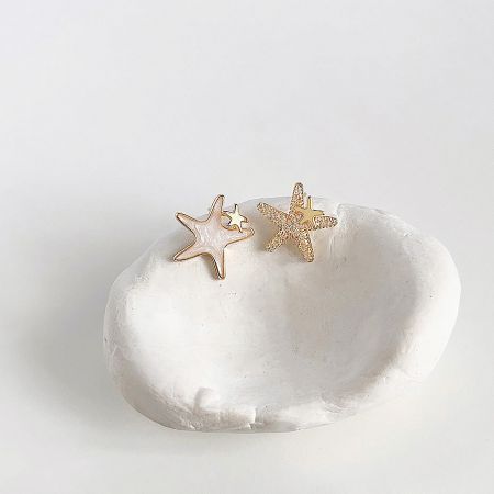 14K Gold Plated Enamel & Zircon Starfish Asymmetrical Stud Earrings (Set of 5 Pairs) - A pair of 14k gold-plated asymmetrical star stud earrings from a set of five, one with a textured gold finish and the other with white enamel, displayed on a decorative stone.