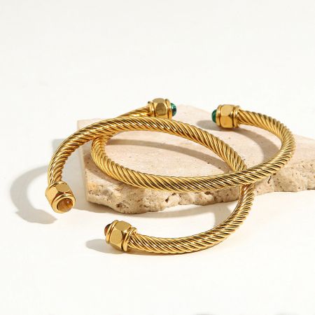 Three stacked stainless steel cuff bangles with twisted design and gemstone ends, displayed on a beige stone.