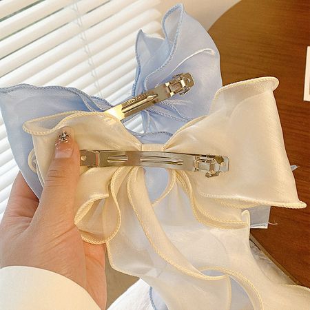 A product shot showcasing two hair bows, one light blue and one white, made of silk or satin with gold hair clips.