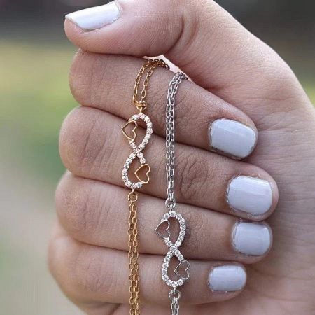 A hand holding the sterling silver infinity heart bracelet, available in silver and gold tones.