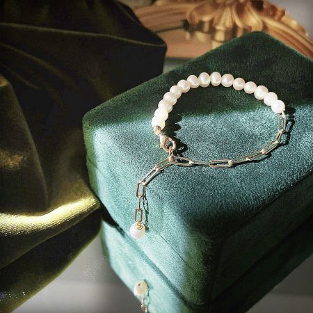 A delicate pearl bracelet with a gold paper clip chain and charm, displayed inside a green velvet jewelry box.