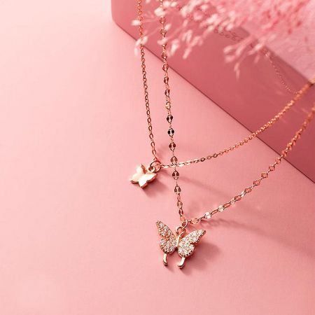 Detailed view of two rose gold butterfly necklaces, one plain and one stone-adorned, on a pink background.