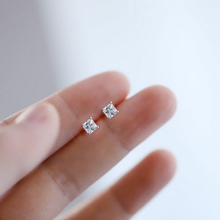 A pair of mini square cubic zirconia stud earrings held between fingers to show scale.