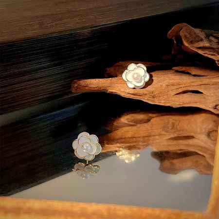 A pair of white and gold Camellia flower earrings with pearl centers displayed on natural driftwood.