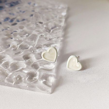 A pair of white heart-shaped enamel stud earrings shown on a clear textured sheet.