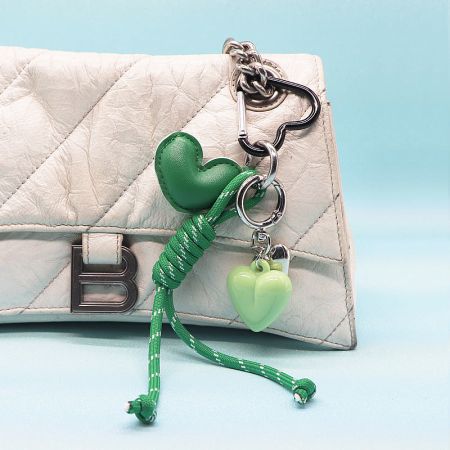 A green PU leather heart keychain with a matching green rope and a silver heart charm, displayed on a white bag.