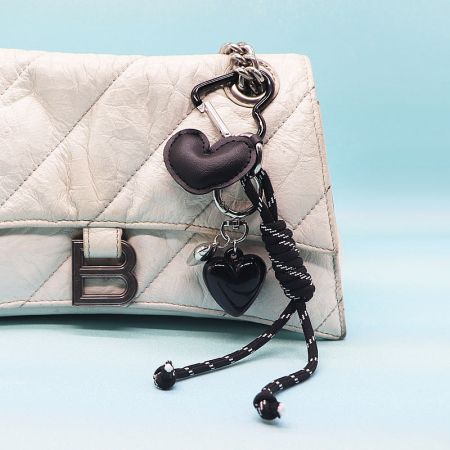 A chic white quilted handbag featuring a black PU leather heart keychain with a bell accent.