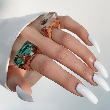 A model's hand wearing two wood resin rings, one with a green crystal and one with a clear crystal.