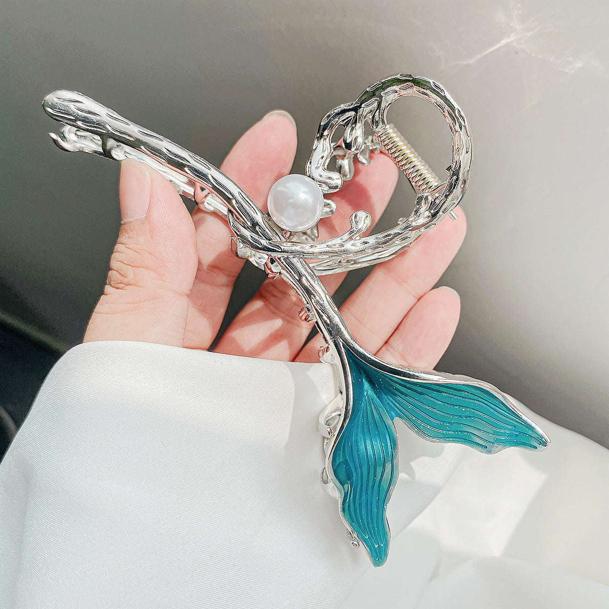 A close-up of a metal alloy hair clip designed as a mermaid tail, featuring a silver finish, a white pearl accent, and a vibrant turquoise tail.