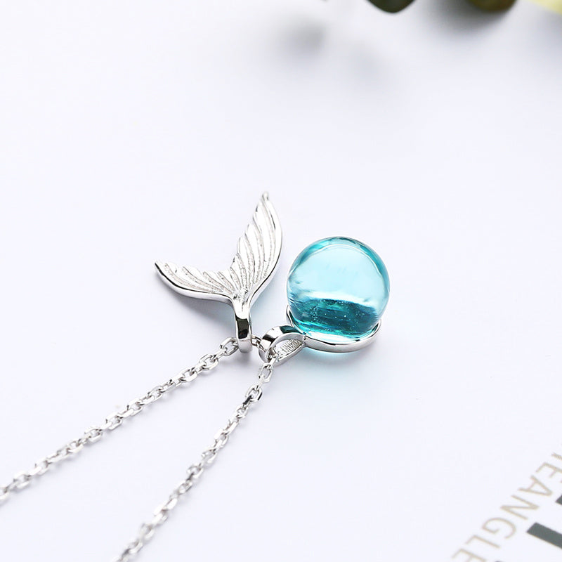 A close-up of a 925 sterling silver necklace featuring a delicate whale tail pendant with a shimmering light blue moonstone bead.