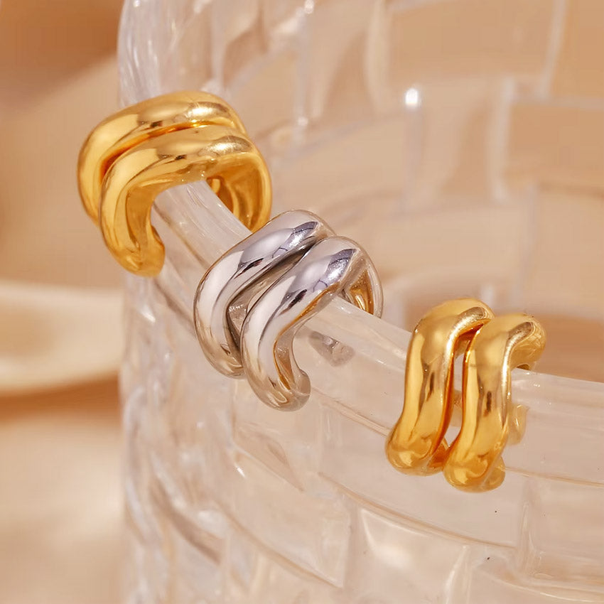 A set of three classic stainless steel cartilage ear cuff earrings in gold and silver finishes, displayed on a glass object.