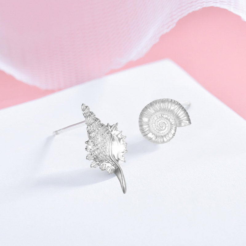A set of five pairs of 925 sterling silver asymmetrical sea shell and conch stud earrings displayed on a pink and white background.