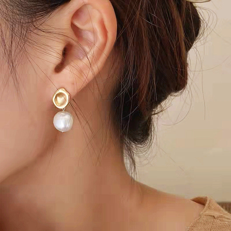 A close-up of a 14k gold-plated baroque freshwater pearl earring with a matte gold heart-shaped stud.