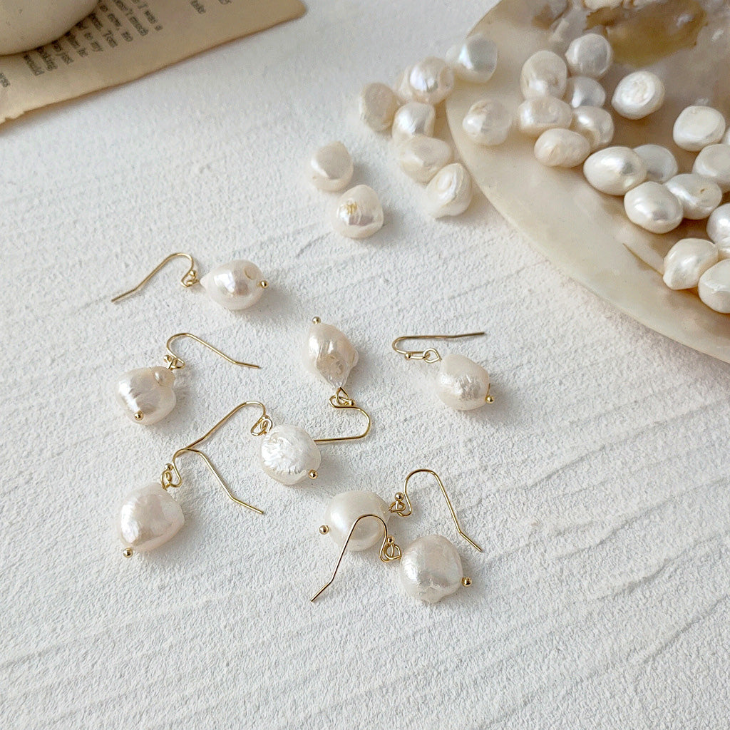 A collection of Baroque irregular freshwater pearl drop earrings with gold hooks, displayed alongside loose pearls on a clean white surface, showcasing the set of five pairs.