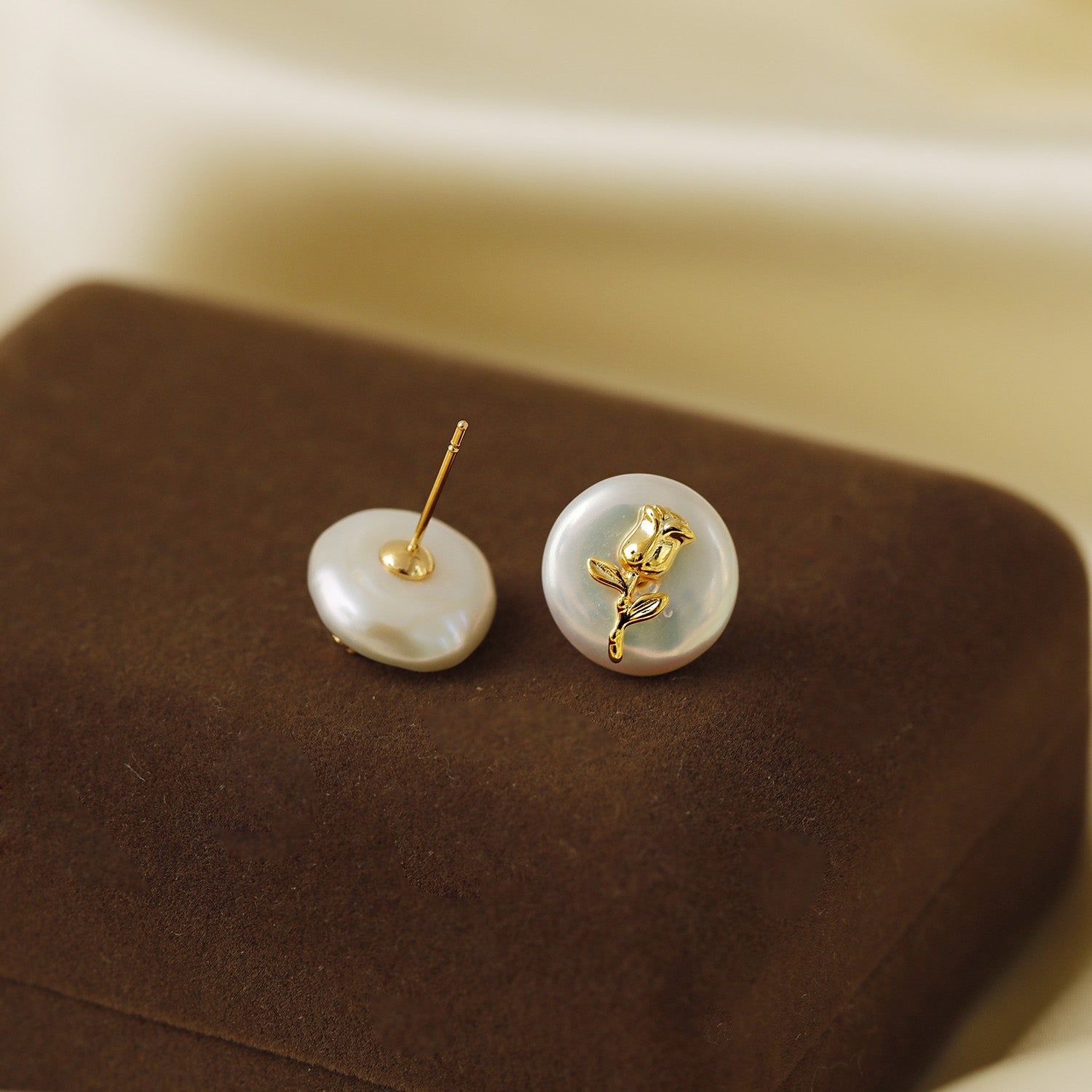 A pair of Baroque pearl stud earrings with a delicate gold flower design, elegantly displayed on a brown velvet stand.