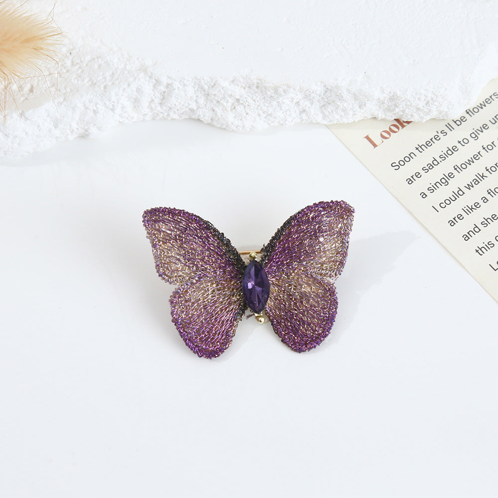 A detailed close-up of the purple hand-embroidered fabric butterfly brooch, featuring intricate wing details and a sparkling central crystal.