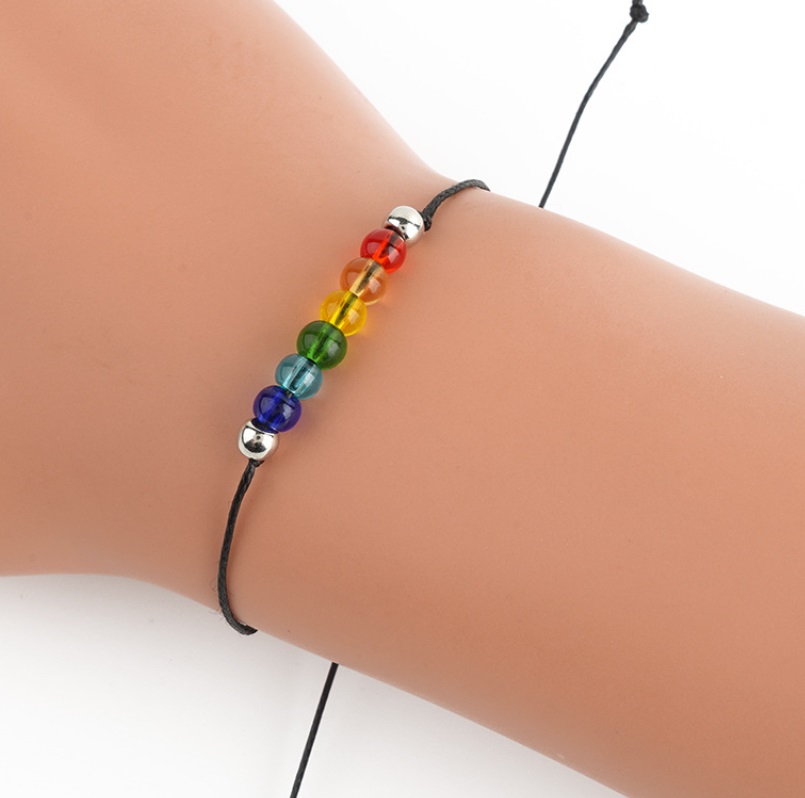 A close-up of the handmade rainbow LGBT rope bracelet being worn, highlighting its colorful acrylic beads and adjustable black cord.