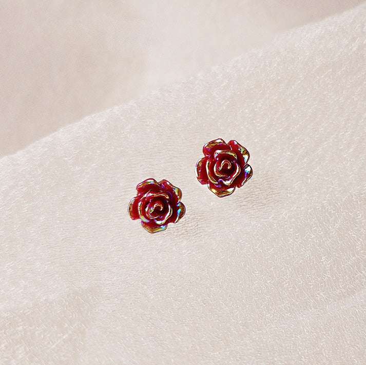 A pair of vibrant red, 3D resin rose stud earrings displayed on a neutral textured background, highlighting their detailed craftsmanship.
