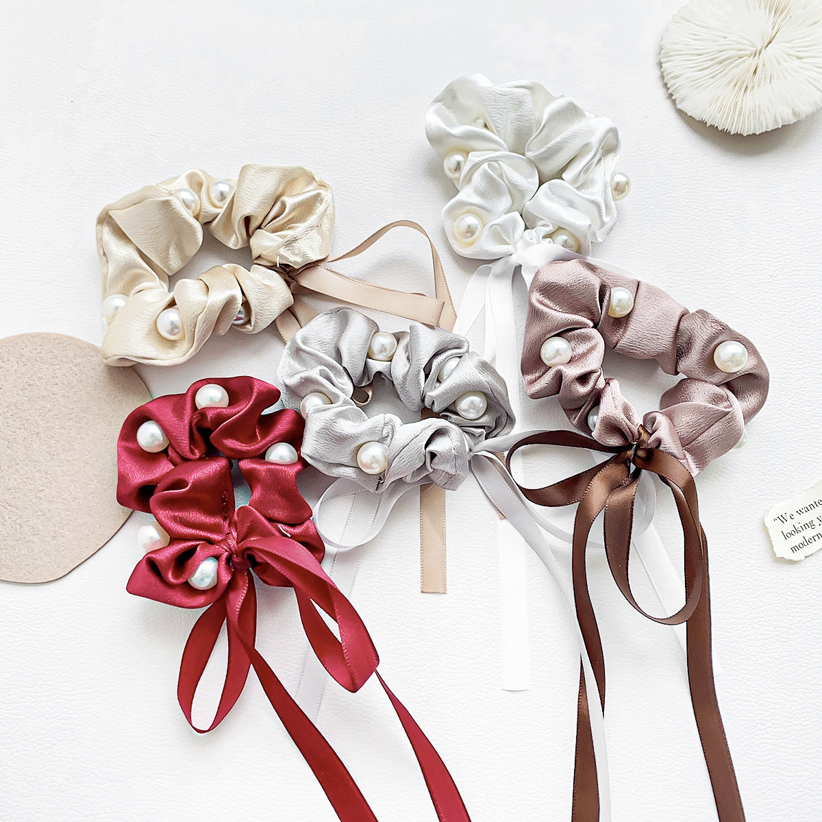 A collection of vintage-style silk scrunchies in various colors, including pink, beige, and gray, adorned with delicate pearls and ribbons.