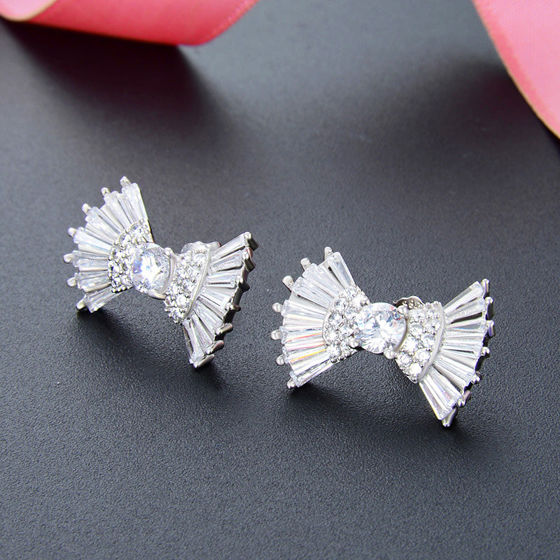 A pair of 925 sterling silver ribbon bow stud earrings adorned with sparkling clear zirconia crystals, displayed on a dark gray background.