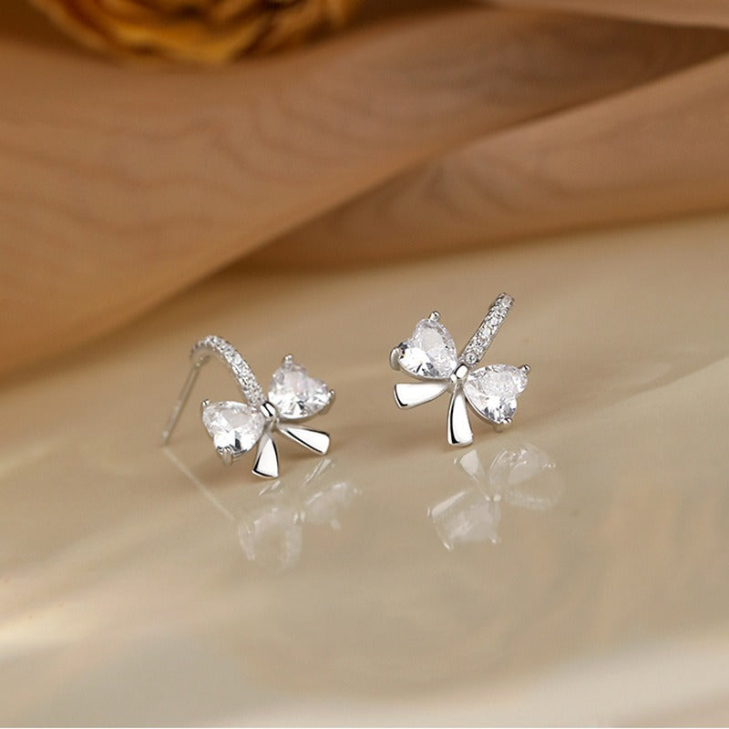 A pair of sterling silver bow stud earrings with sparkling heart-cut cubic zirconia stones, presented on a neutral background.