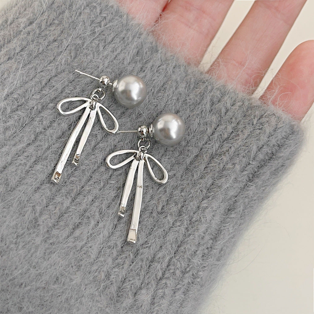 A pair of silver-plated drop earrings with grey pearl studs and delicate bowknot charms with long tassels, displayed on a textured grey surface.