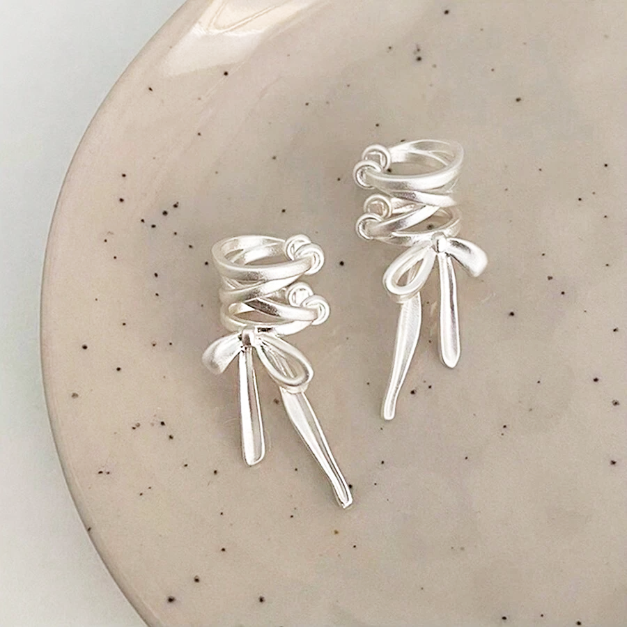 A pair of 925 sterling silver matte bowknot ear cuff earrings with a unique coiled design, displayed on a decorative plate.
